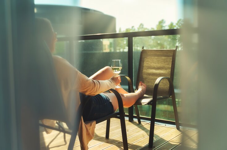 Eine Person sitzt entspannt auf einem Balkonstuhl, hält ein Glas Weißwein in der Hand und genießt die Sonne, mit ausgestreckten Beinen auf einem zweiten Stuhl.