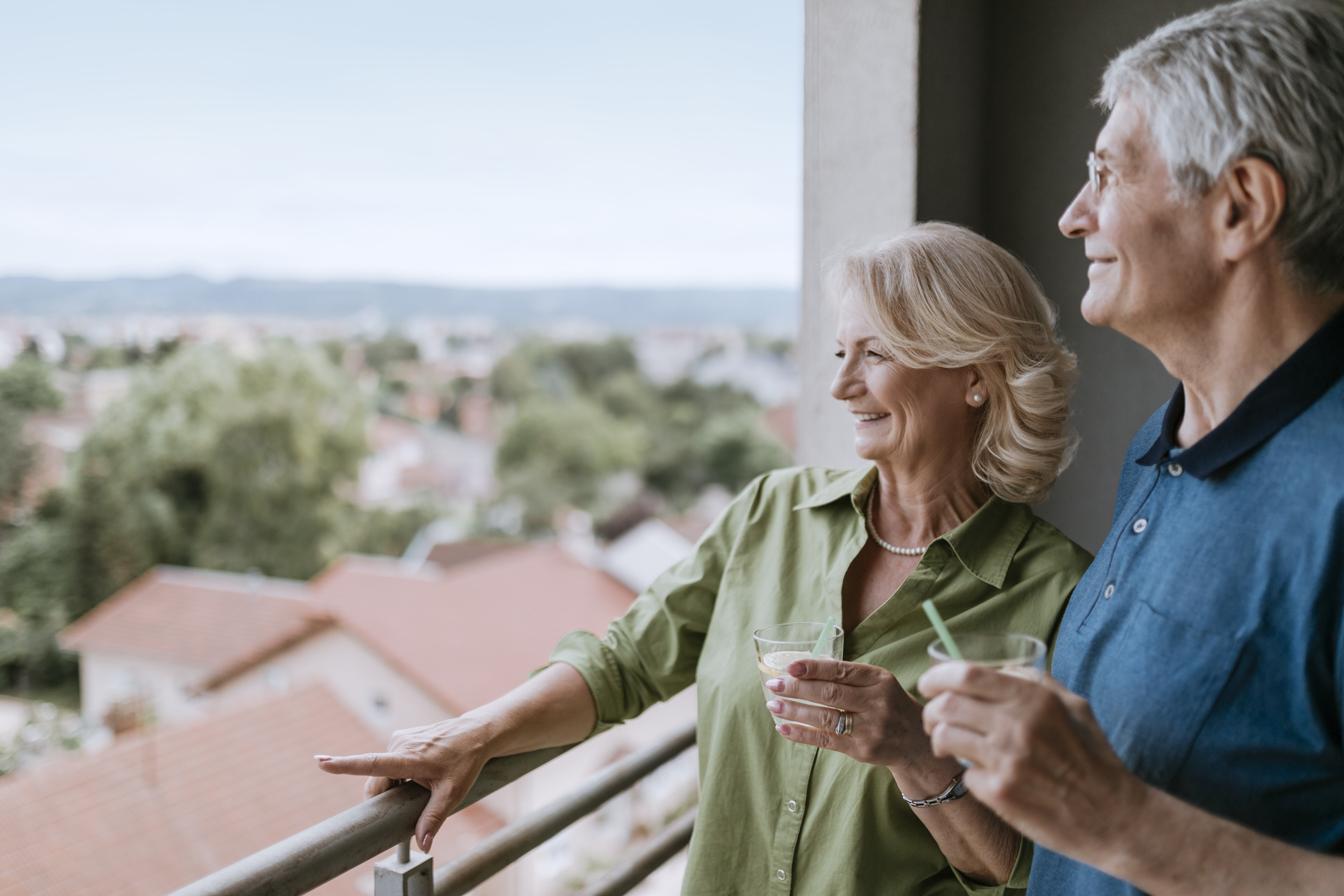 Vertrauen & Sicherheit Ein älteres Paar steht lächelnd auf einem Balkon und genießt gemeinsam ein Getränk, während sie die Aussicht auf eine ruhige Wohngegend mit roten Dächern und grüner Umgebung betrachten.